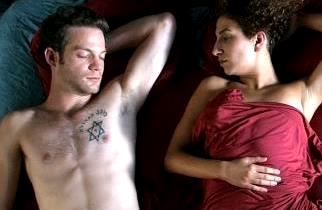 A young man without a shirt sleeps next to a young woman who is covered by a blanket.